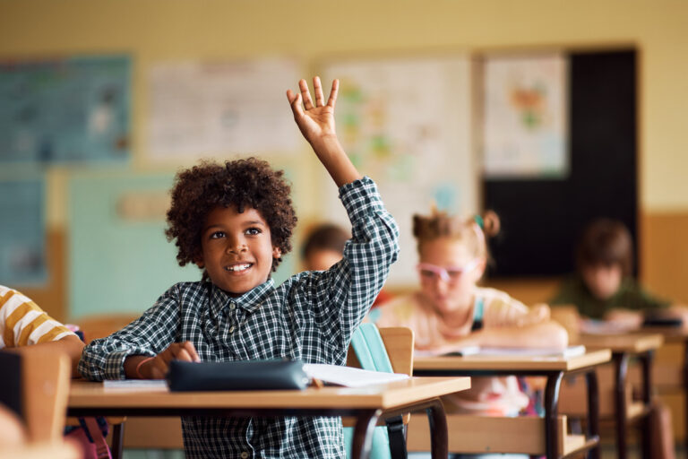 happy male student raising hand to answer question in elementary classroom